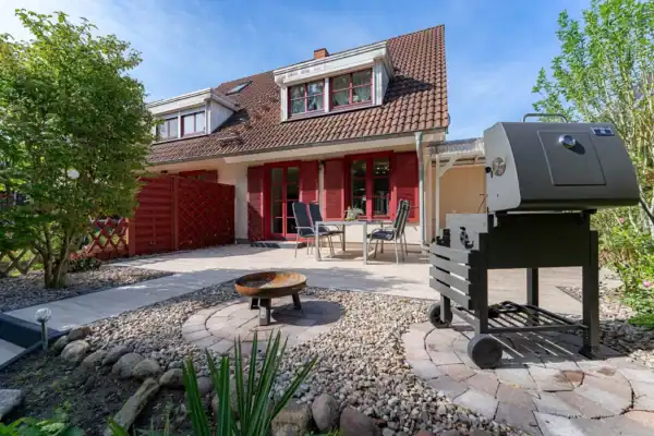 Außenansicht, vorne rechts Grill, vorne mitte Feuerschale, vorne links Treppe zum Garten, hinten links Terrassentür mit roten Fensterläden, hinten mitte Gartentisch mit Stühlen