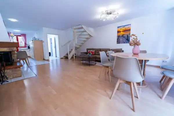 Wohnzimmer, links Kaminofen und dahinter offene Küche, rechts Esstisch mit 6 Stühlen, hinten rechts Sitzecke, links daneben Treppe nach oben und Tür zum Flur