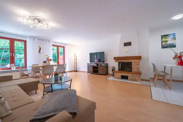 Wohnzimmer, vorne links Sitzecke mit Couchtisch, hinten links Esstisch mit 6 Stühlen, hinten mitte TV und Kaminofen, hinten rechts Tisch mit 4 Stühlen